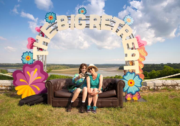 two woman sat on a sofa under a festival sign The Big Retreat, with views of the an estuary in the background.