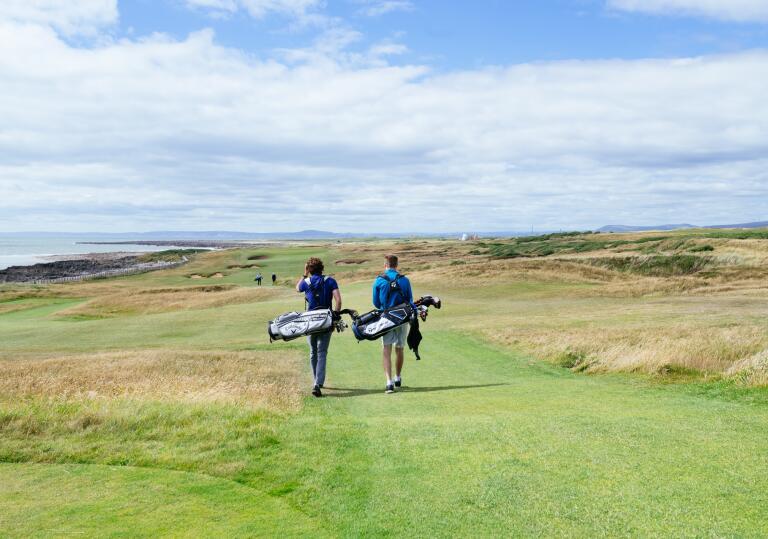 Two people with golf bags walking down a wide pathway on a coastal golf course.