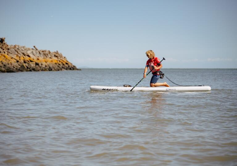 A woman kneeling down on a stand up paddleboard on the sea, using a paddle.