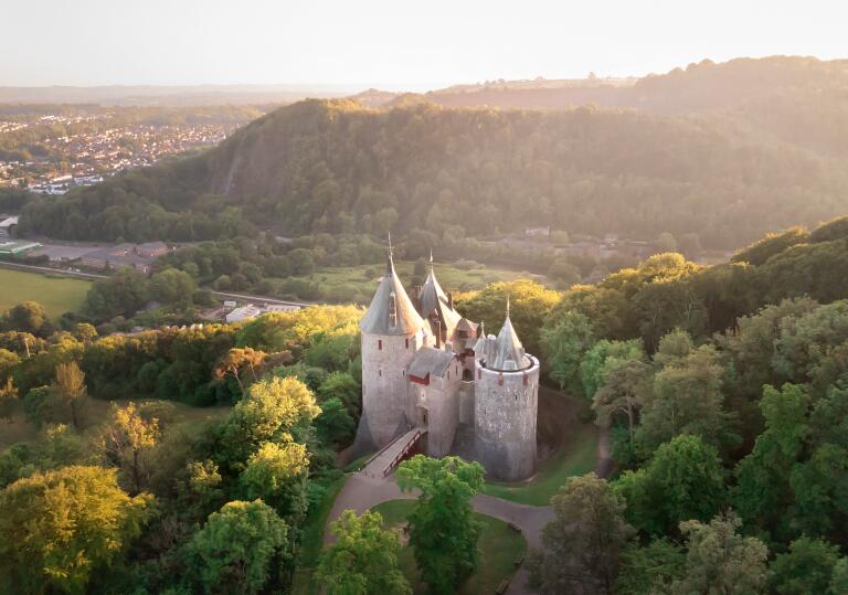 Aerial shot of a fairy tale style castle with pointy turrets and a drawbridge, set amoungst the trees of a woodland. 
