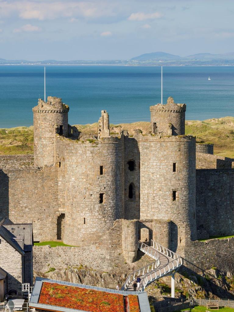A view of Harlech Castle and its white walkway leading to the entrance with the sea and golf course beyond, 