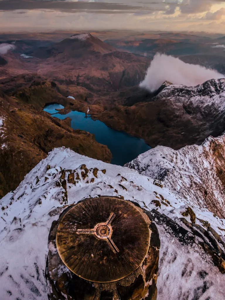A waymarker at the top of a snow-topped mountain, with a view down over a blue lake