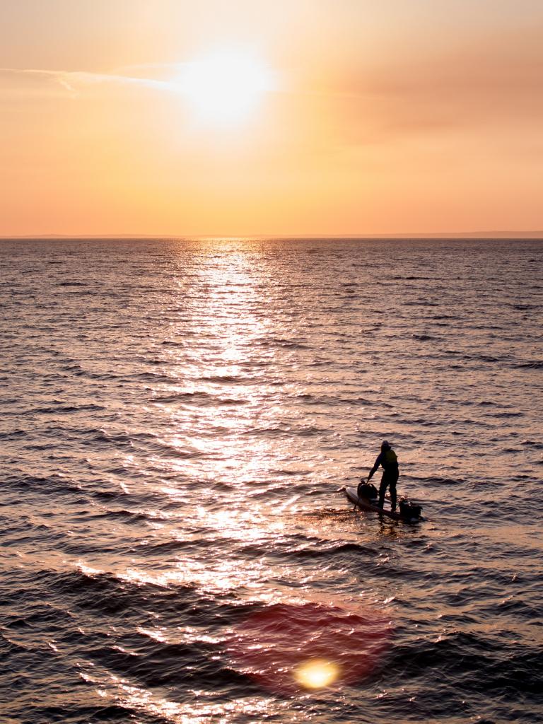 A person paddleboarding on the sea with a sunset behind.