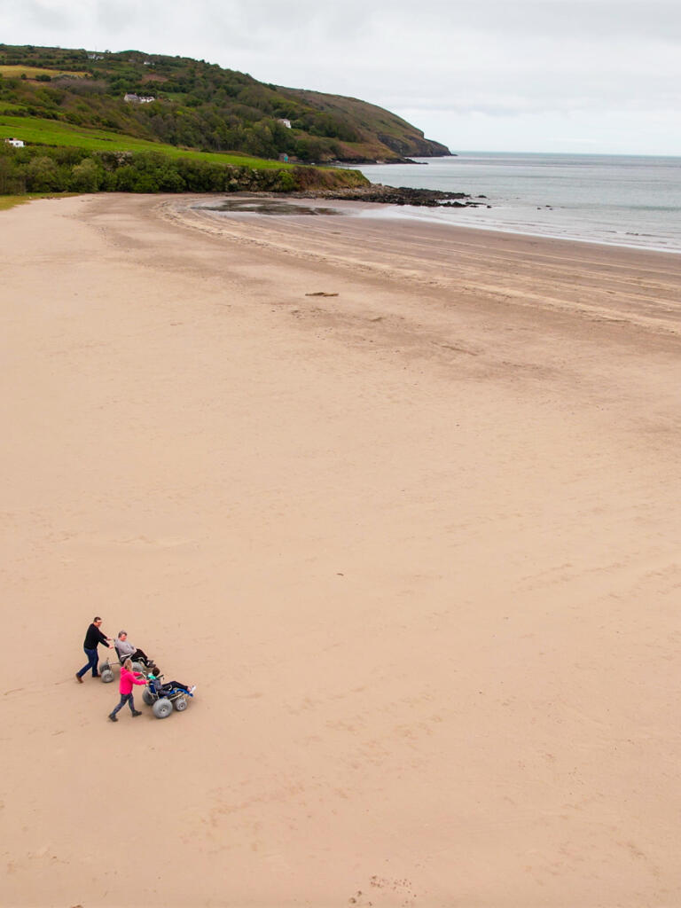 A drone shot of a wide sandy beach with two people using beach wheelchairs.