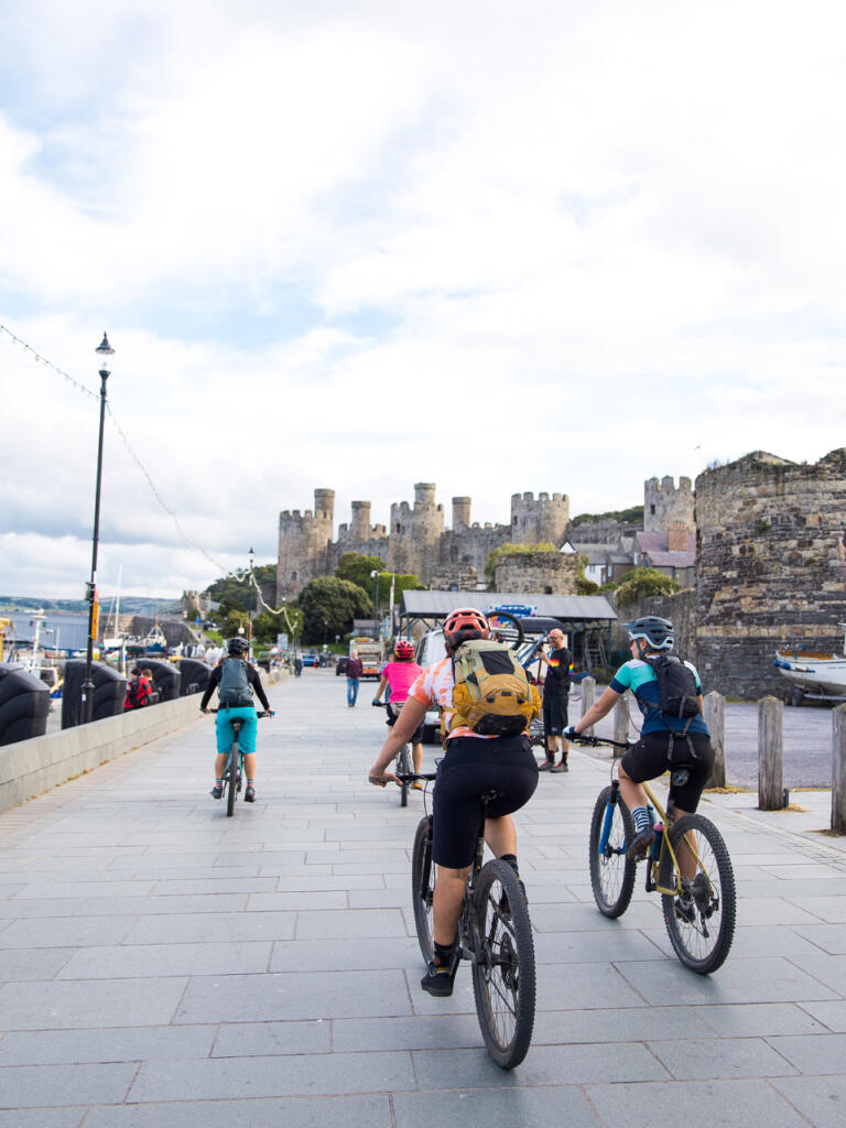 A group of cyclists on mountain bikes on a wide promenade, approaching a large castle.