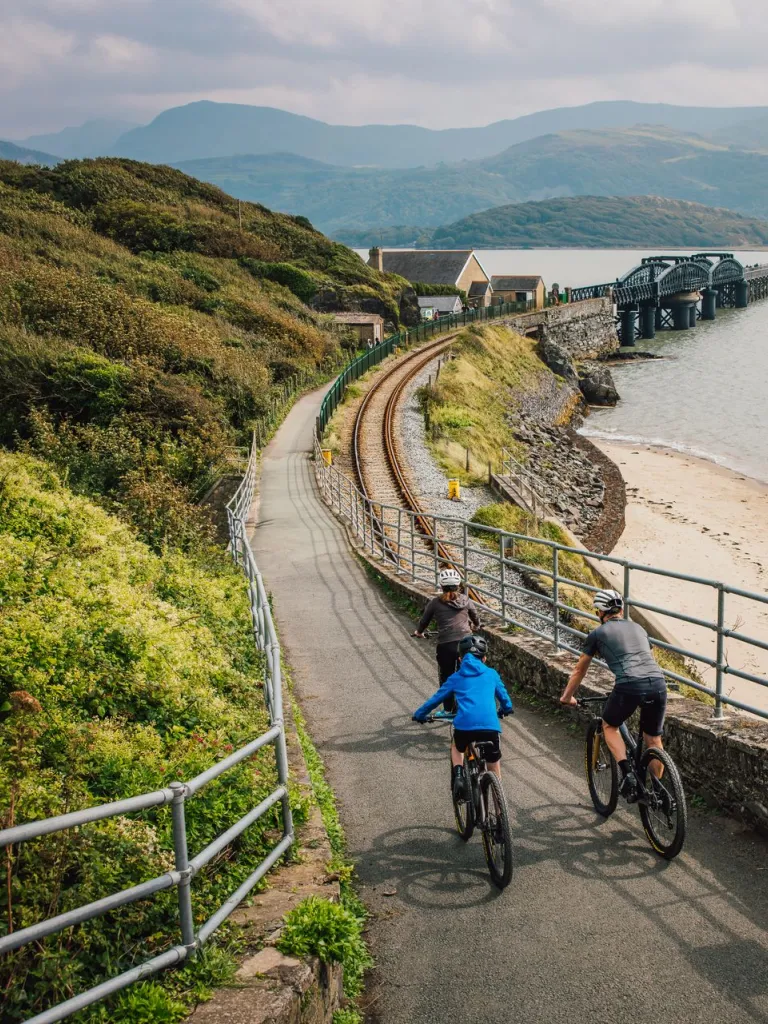Three cyclists on a cycle path approaching a wooden railway bridge over a wide estuary.
