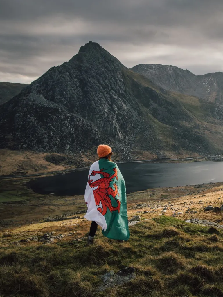 A person wearing a Welsh flag as a cloak overlooking a lake and a range of mountains.