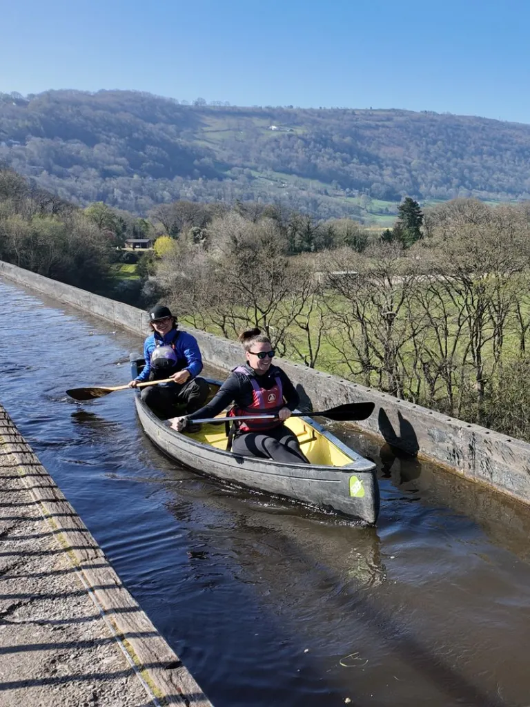 Canoes being paddled across a narrow canal aqueduct, with a steep drop on one side.