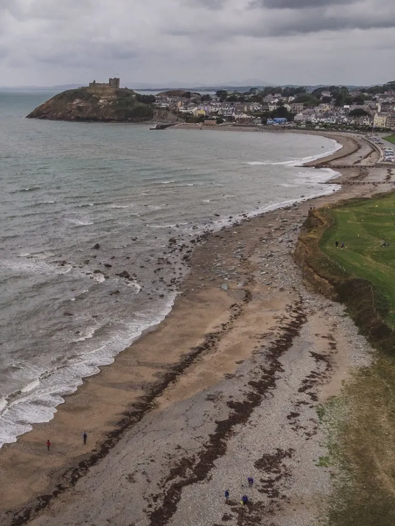 A long pebbly beach in a bay, looking towards a ruined castle and a town.