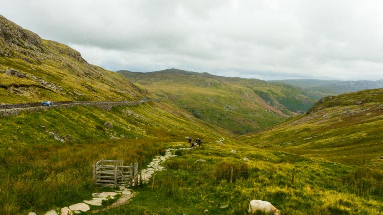 Walkers on a path through a valley of mountains, with sheep grazing.