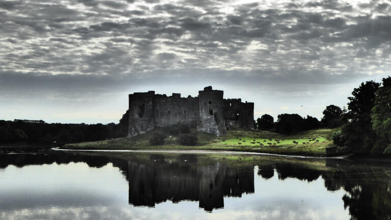 view of Carew castle on the banks of a river with clouds above. The castle and clouds and reflected in the river like a mirror.s