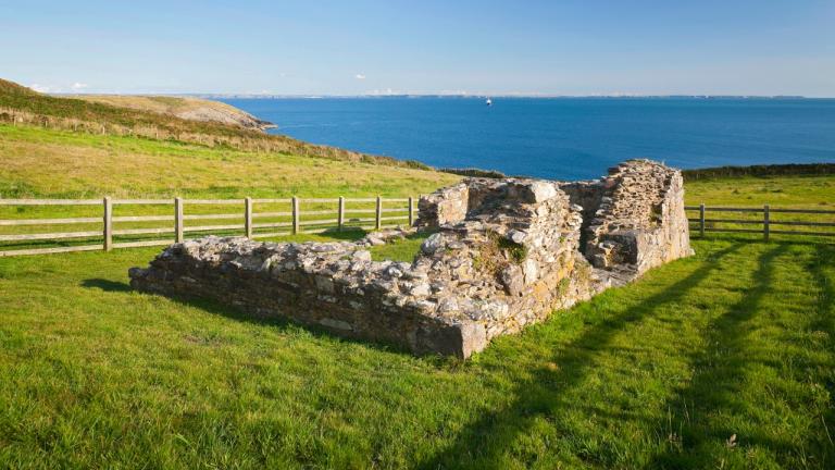 The ruins of St Non's Chapel with a wooden fence surrounding it overlooking the sea.