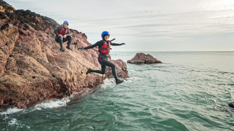 A lady jumping off a rock into the sea wearing safety gear with a friend looking on.