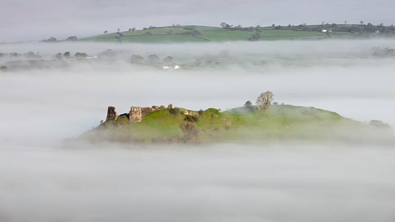 Fernsicht auf eine Burgruine auf einer Hügelkuppe, die in tiefhängende Wolken gehüllt ist.