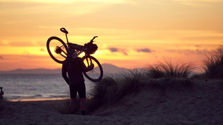 The adventurer, Richard Parks, carrying a mountain bike on the beach in Barmouth at sunset.