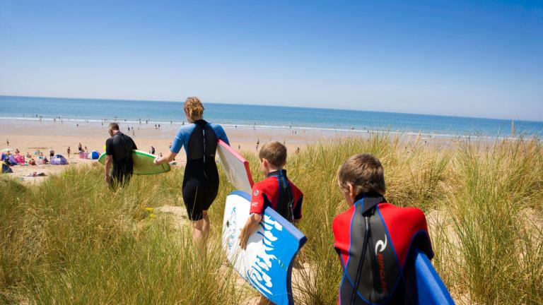 Family walking along sand dunes to the sea with surf boards