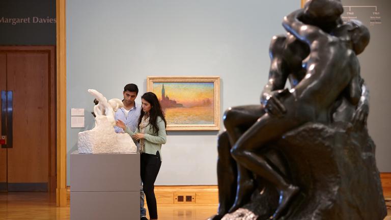 A couple looking at a small statue, with a painting behind them, and a large sculpture to the side in the foreground