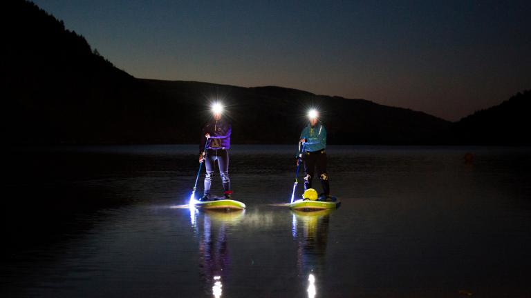 Two people stand up paddleboarding at night on a lake.