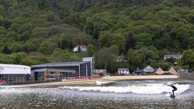 Surfing in foreground and countryside and buildings in background at Surf Snowdonia