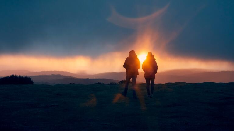 A silhouetted couple watching a low orange sun from a hill top.