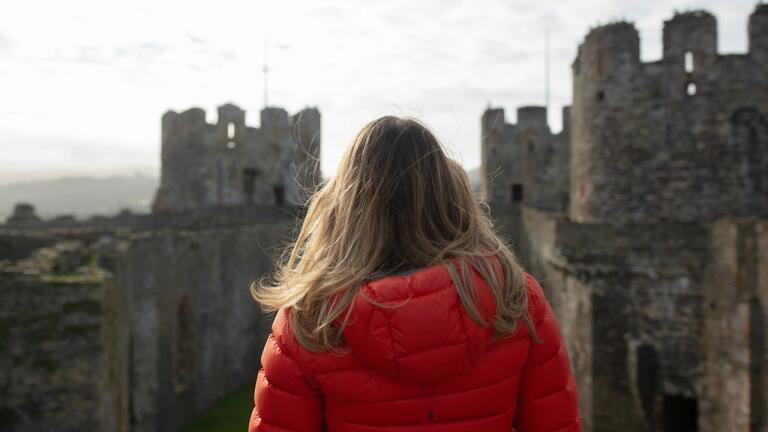 A girl in a bright orange coat with her back to the camera looks out across the towers of a castle