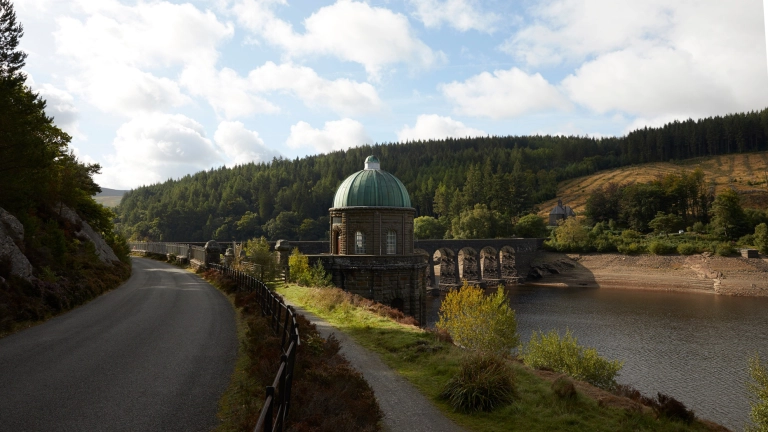 Ein historischer Steindamm mit Kuppelgebäude neben einem Fußweg und einer schmalen Straße.
