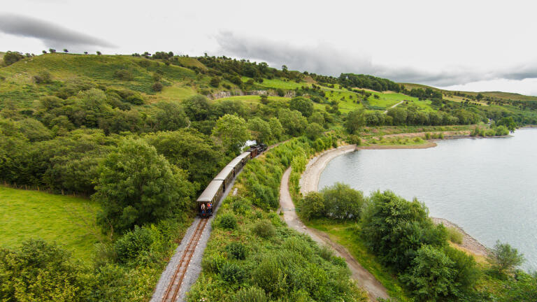 A narrow gauge steam hauled train next to a reservoir.