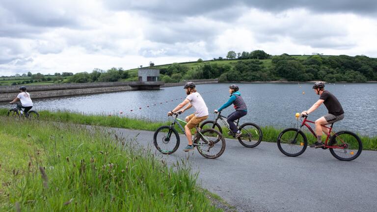 A group of four cyclists on a smooth, level path by a reservoir.