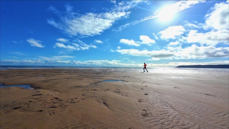A man in tshirt and shorts running along a wide sandy beach, under blue skies.