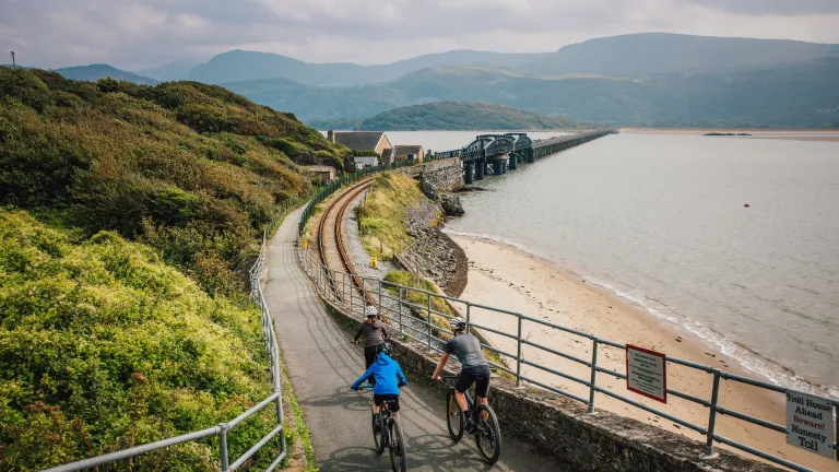 Three cyclists on a cycle path approaching a wooden railway bridge over a wide estuary.