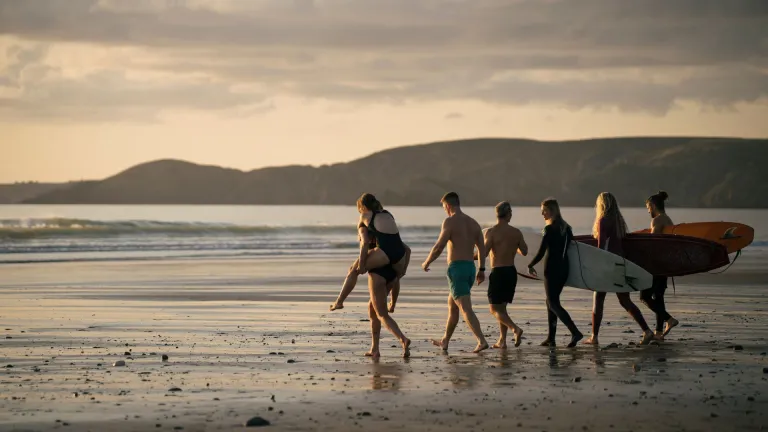 Eine Gruppe von Freunden in Badeanzügen, die mit Surfbrettern ausgerüstet am Strand entlang Richtung Meer laufen.