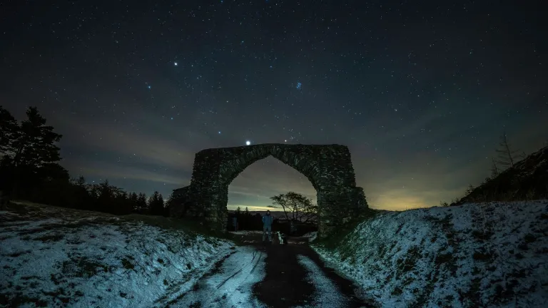 A stone built archway over a snowy narrow road at night. The stars are bright in thedark skies.