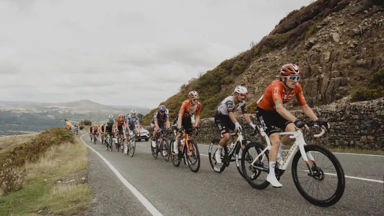 Peloton riding the 2025 Tour of Britain on the Tumble climb out of Blaenavon, winding along a rural road beside a stone wall in the Welsh countryside.