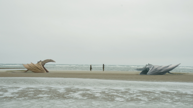 Two dragons on a wide sandy beach with two people standing in between them.