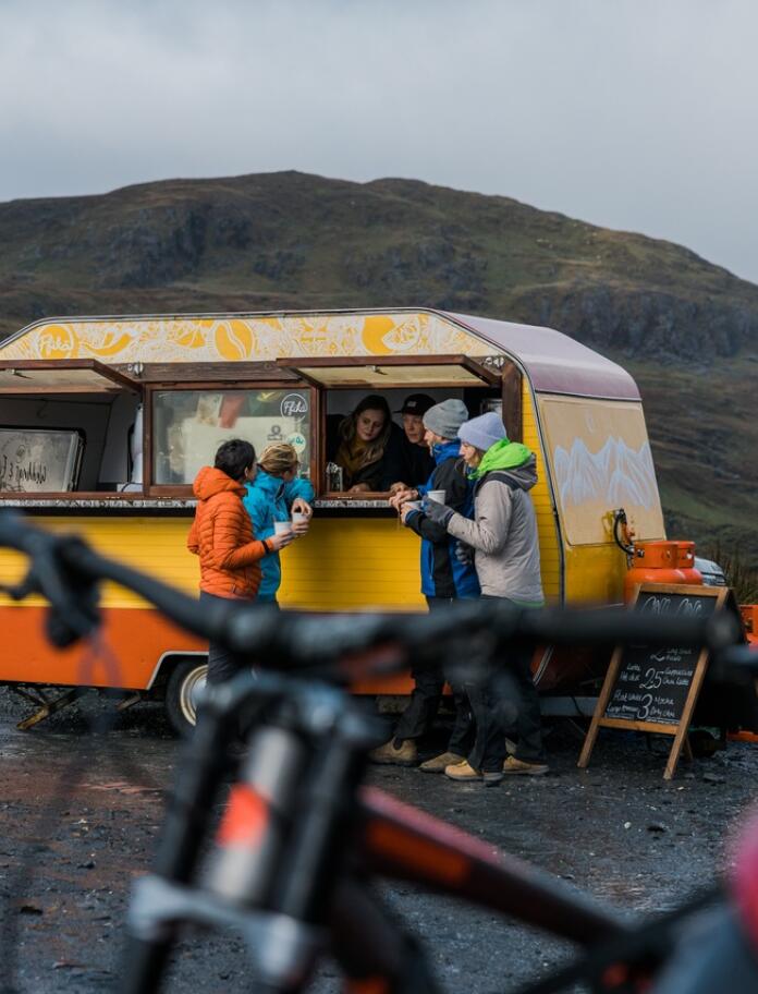 Vier Menschen die sich bei Fikka stärken, einem gelben Wohnwagen, der in einen Kaffee-Stand verwandelt wurde. Berge im Hintergrund und ein Mountainbike im Vordergrund.