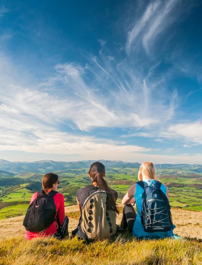 Three female walkers sat on the ground looking out across valleys and hills.