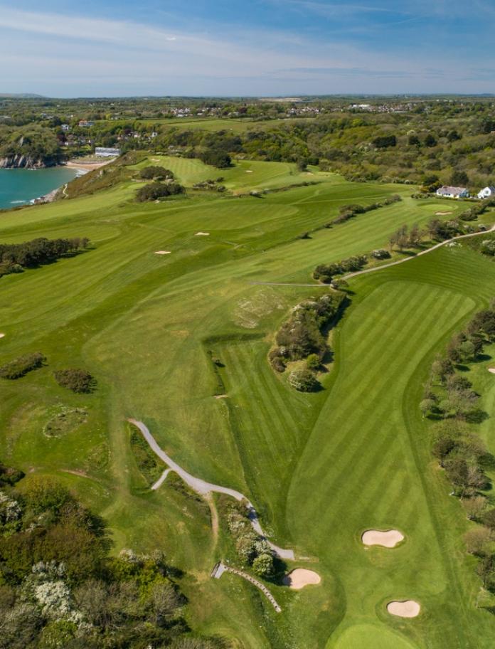 An aerial shot of a golf course and the blue waters of a bay alongside it.