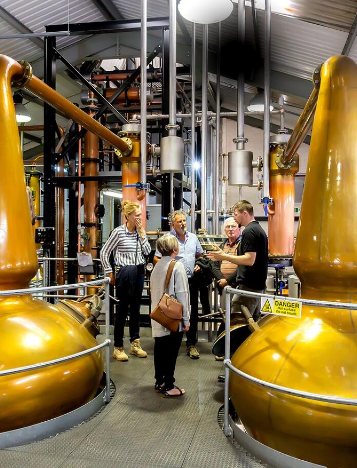 A group of people taking a tour of a distillery, next to two golden vats.