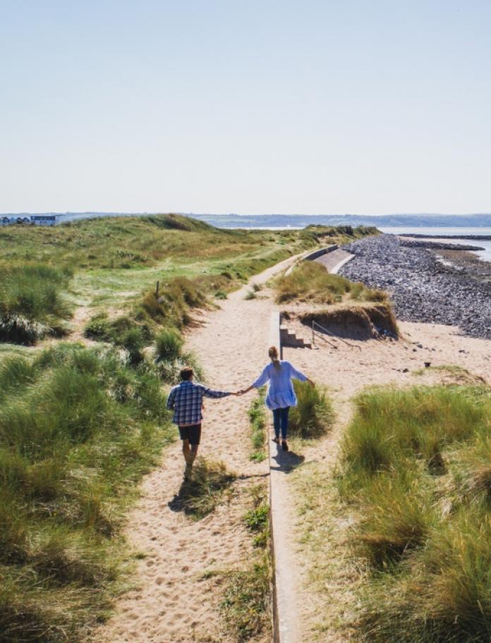 Couple walk hand in hand along path between sand dunes with sea to one side