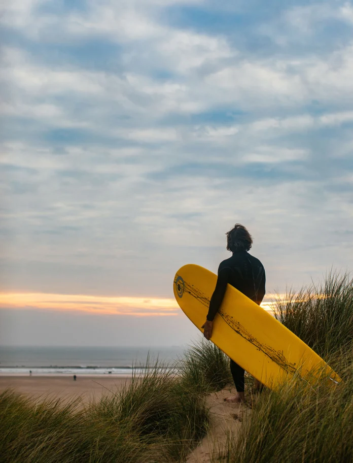 surfer holding surf board with tall grass and sea in background.