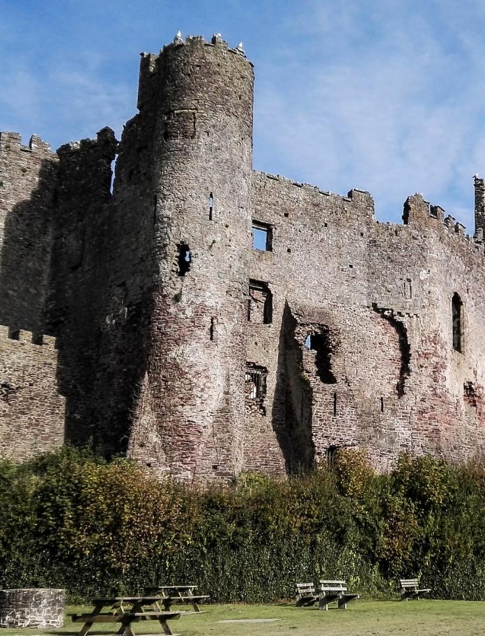An external shot of Laugharne Castle, with people walking up the path next to green lawns.
