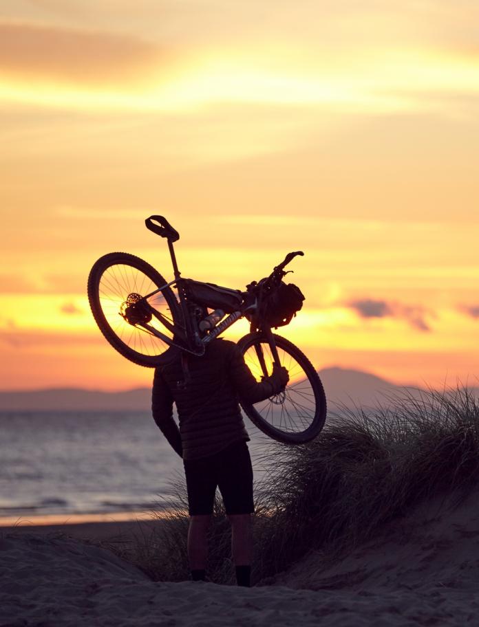 The adventurer, Richard Parks, carrying a mountain bike on the beach in Barmouth at sunset.