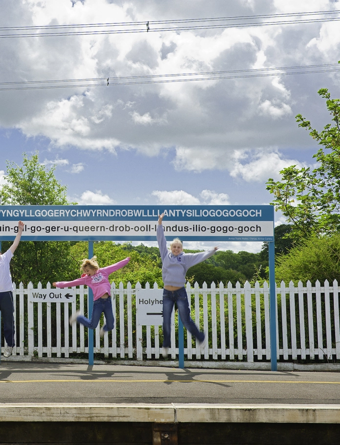 Image of three people in front of the sign for Llanfair­pwllgwyngyll­gogery­chwyrn­drobwll­llan­tysilio­gogo­goch in Anglesey.