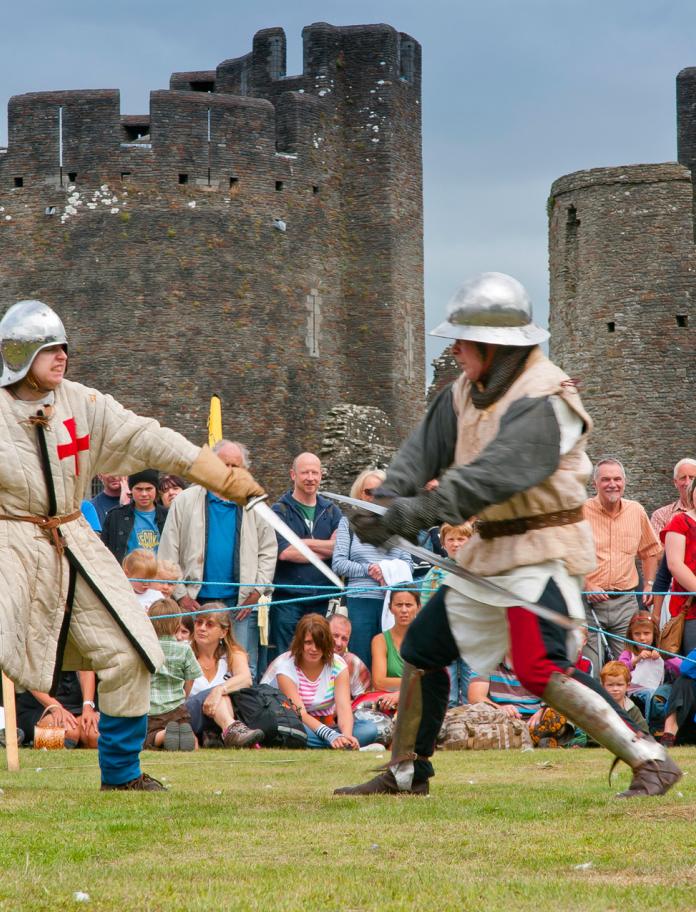 Two men dressed in medieval costumes taking part in a battle re-enactment, with crowd watching by the castle.