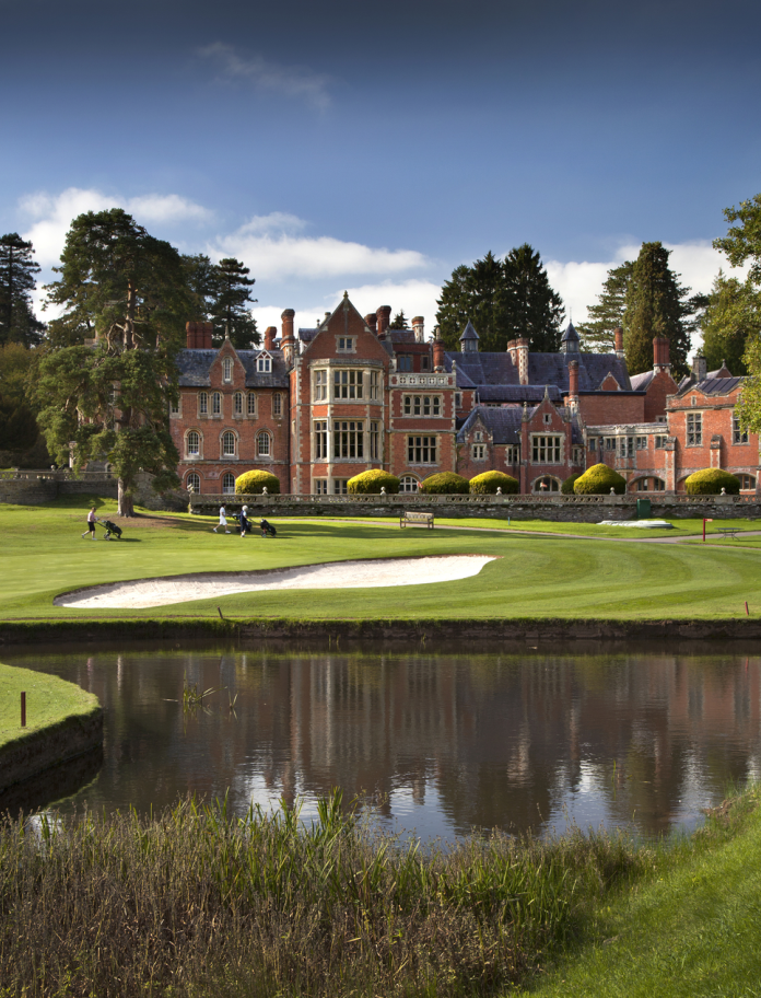 Golfplatz mit einem See im Vordergrund, Bunkern und Herrenhaus im Hintergrund.