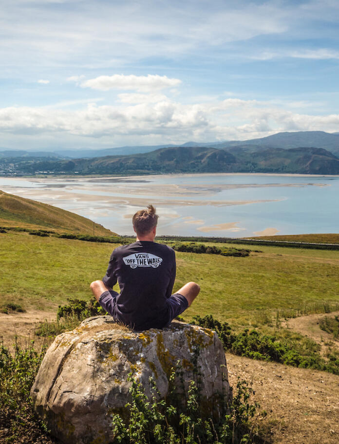 A man sitting on a rock overlooking some countryside and the coast.