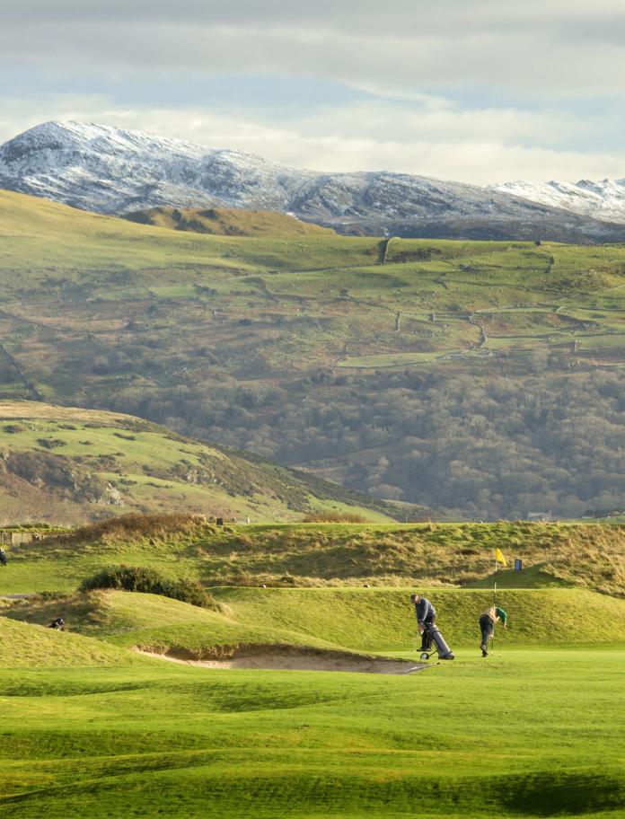 People playing on Porthmadog Golf Club green with snow-capped mountains in the background.