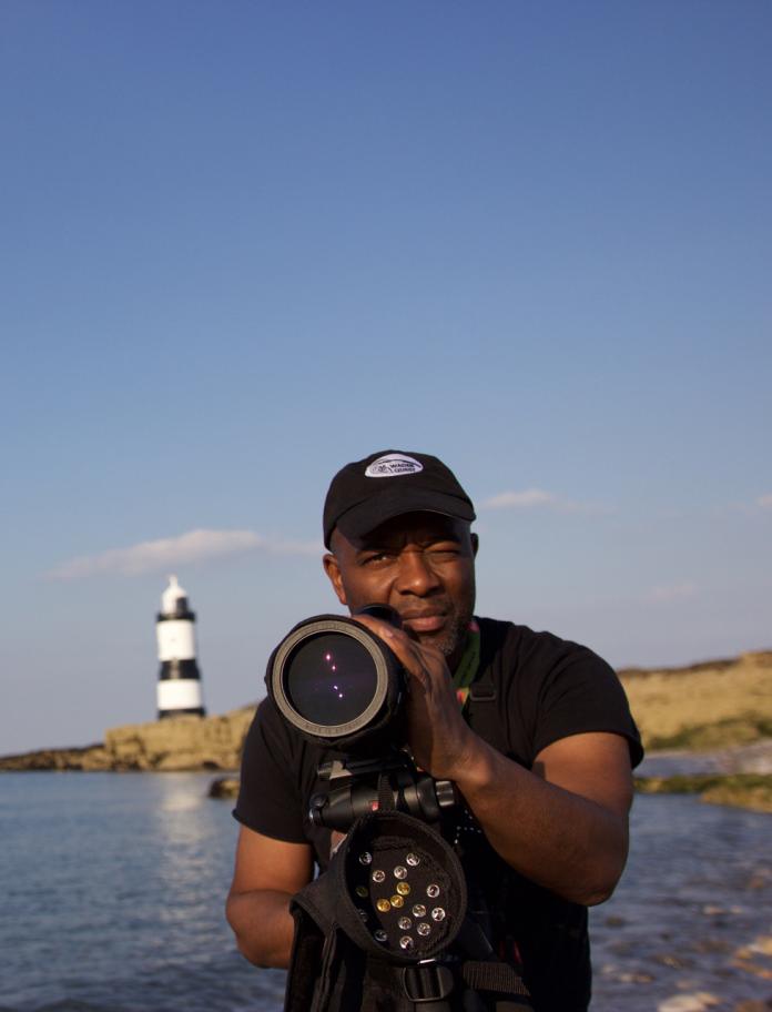 David Lindo holding a camera with a lighthhouse and sea in the background.