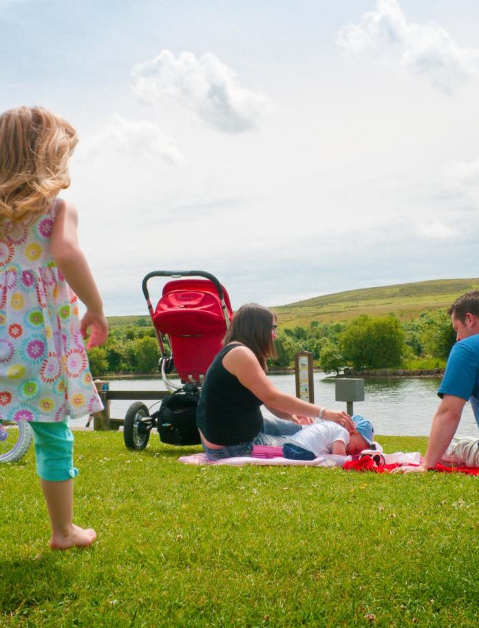 young girl running with parents and baby sat on picnic mat overlooking a lake.