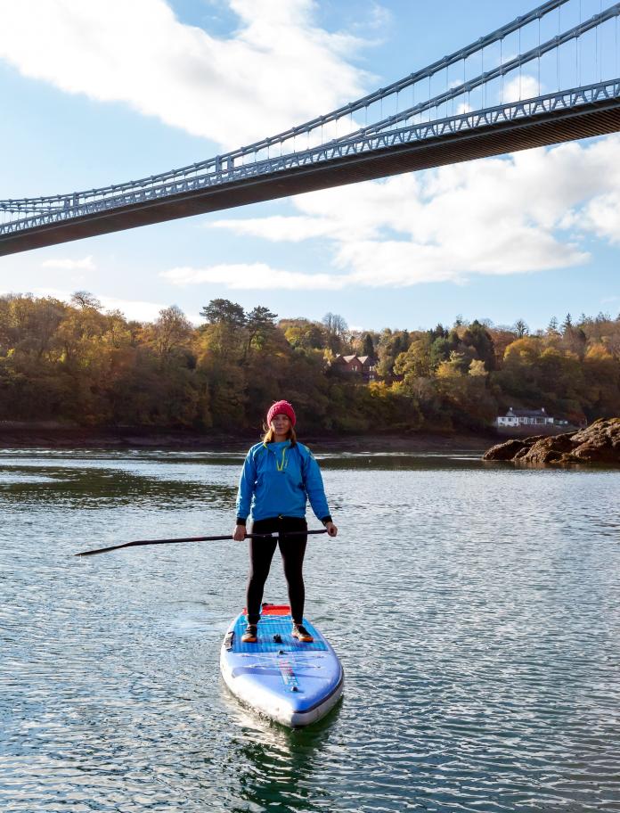 A woman on a paddleboard with the Menai Suspension Bridge in the background.